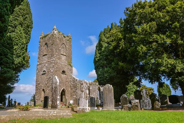 A weathered stone church tower among headstones, framed by tall trees under a blue sky.