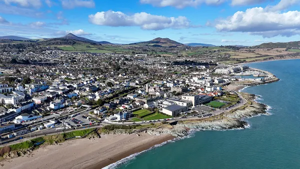 A drone view of a coastal town with a sandy beach and harbour, green hills and mountains behind.
