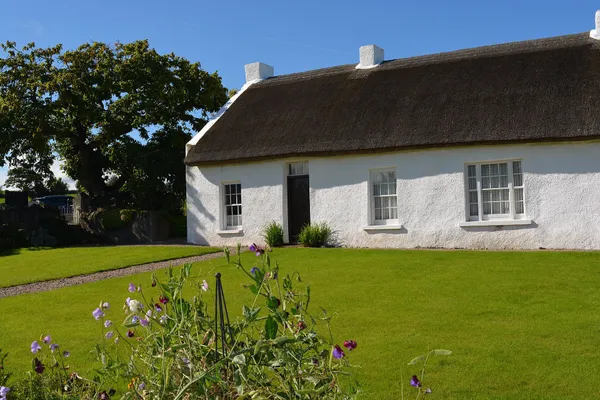 A long white cottage with a thatched roof behind a green lawn and wildflowers under a blue sky.