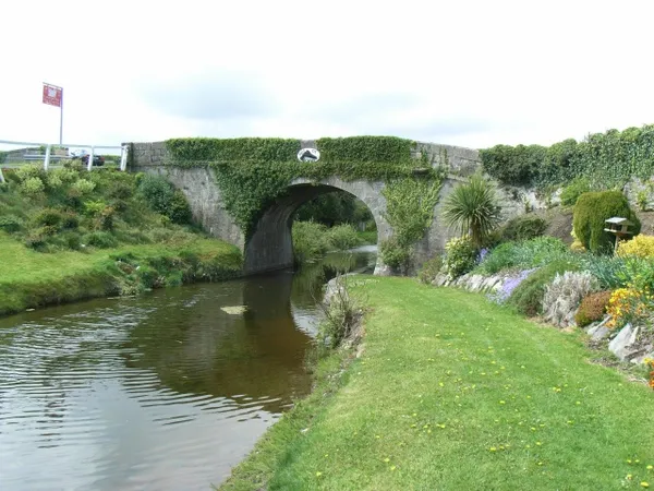 An ivy-covered stone arch bridge over a calm canal, with a grassy towpath and garden flowers.