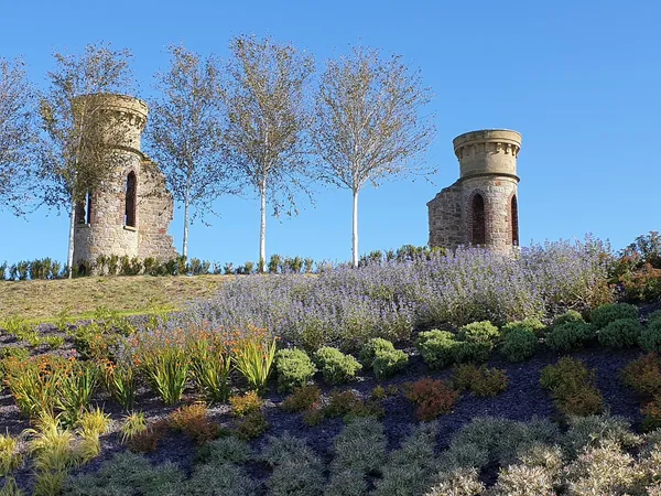 Zwei verfallene Steintürme und schlanke Birken über einem abfallenden Beet aus Lavendel und Sträuchern unter blauem Himmel.