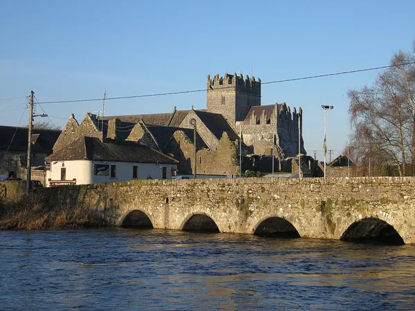Eine mehrbogige Steinbrücke über einen Fluss mit einer grauen Steinabtei und quadratischem Turm dahinter, unter blauem Himmel.