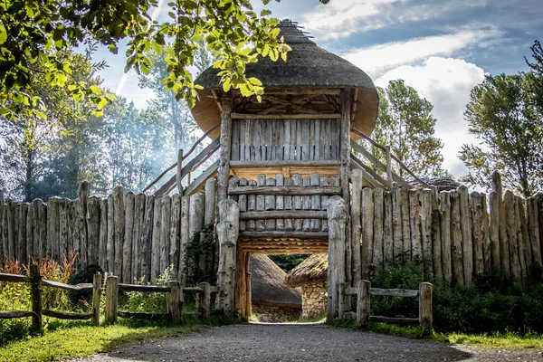 A thatched wooden gatehouse set into a tall log palisade, framed by trees under a bright sky.