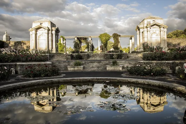 Dos pabellones de piedra unidos por una pérgola, reflejados en un estanque redondo bordeado de parterres de rosas.