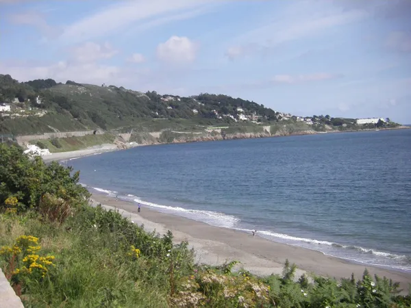 A long curving beach and calm blue sea below green wooded hills dotted with houses.