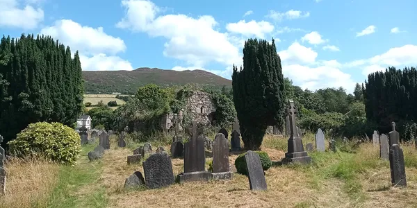 Weathered headstones among dark yews and a ruined stone church, with a green hill behind under a blue sky.