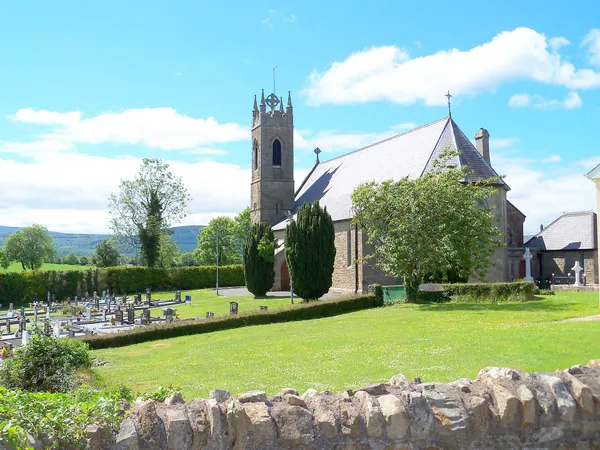 Una iglesia de piedra con torre cuadrada junto a un cementerio y césped verde, con colinas al fondo bajo un cielo azul.