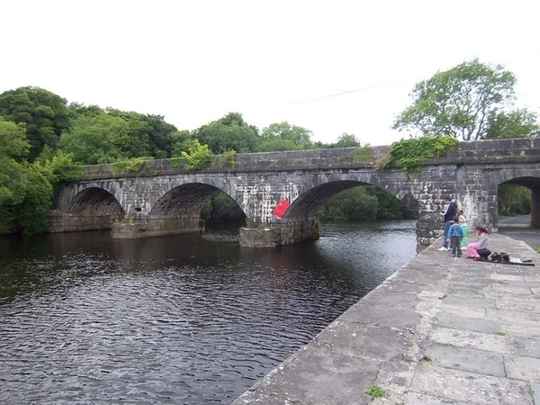 Eine mehrbogige Steinbrücke über einem dunklen Fluss, mit Bäumen dahinter und einem steinernen Kai im Vordergrund.