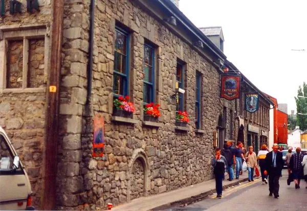 A long stone building with window boxes of red flowers and hanging pub signs, with people walking along the street.