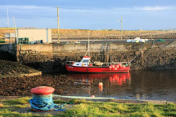 Un piccolo porto con una barca da pesca rossa in acque basse accanto a una banchina in pietra, con un bitta di ormeggio davanti.