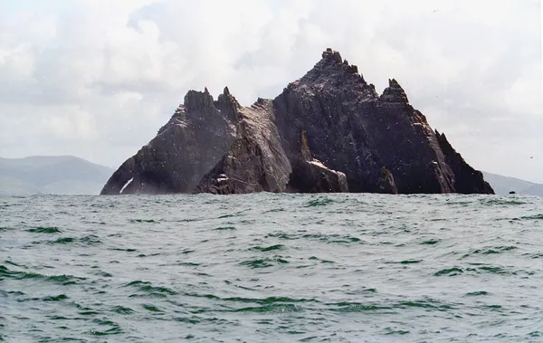 Un'isola rocciosa scura e frastagliata che si innalza ripidamente da un mare grigio-verde mosso, con colline all'orizzonte.