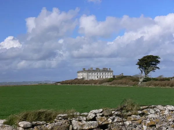 Una gran mansión pálida en el horizonte más allá de campos verdes y un muro de piedra, con un árbol azotado por el viento a su lado.