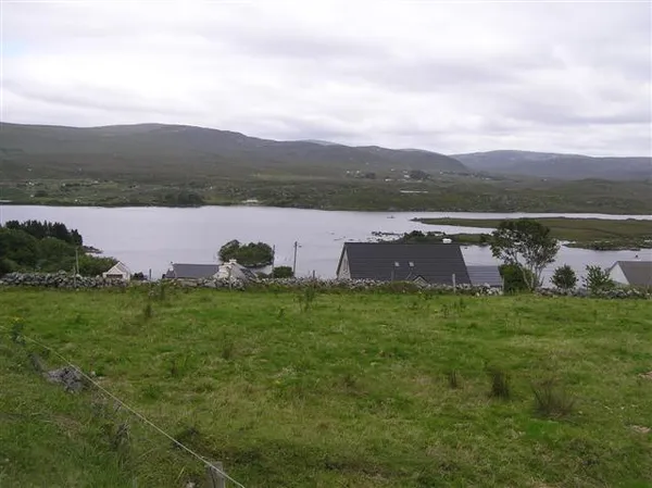 Un campo de hierba frente a una hilera de casas, con un lago y colinas bajas detrás bajo un cielo nublado.