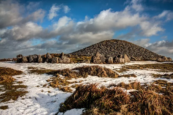 A large stone cairn ringed by kerbstones on a hilltop, with patches of snow and a blue sky with clouds.