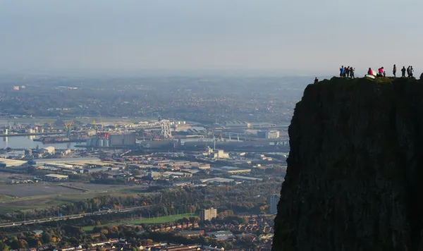 A dark cliff edge with small figures on top, overlooking a wide city and harbour below.