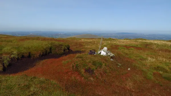A small stone cairn on a grassy, boggy hilltop with a wide view to distant hills.