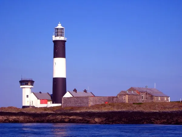 Un phare à bandes noires et blanches à côté de bâtiments en pierre bas et d'une tour blanche plus petite sur une île rocheuse.