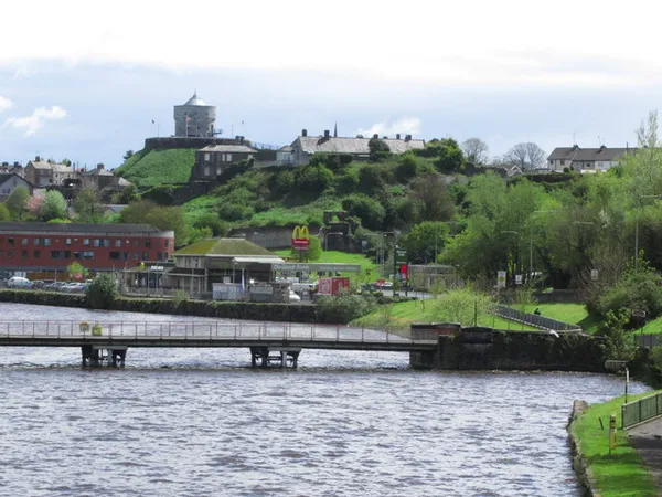 A round stone tower on a green hill above town buildings, seen across a river with a footbridge.