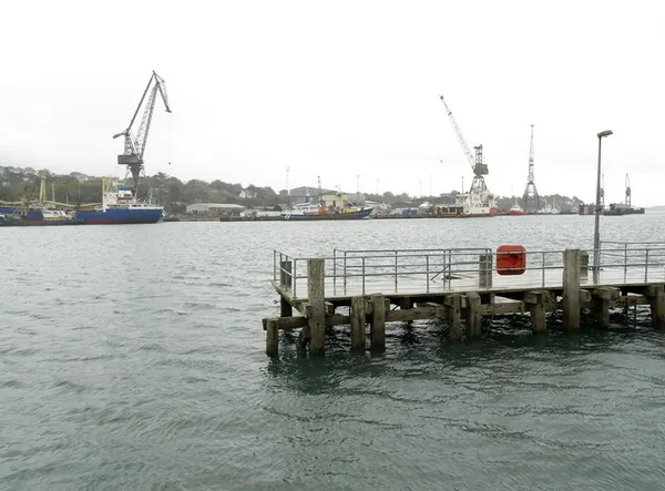 Un piccolo molo di legno che si protende in acque grigie del porto, con gru e navi sulla riva opposta.