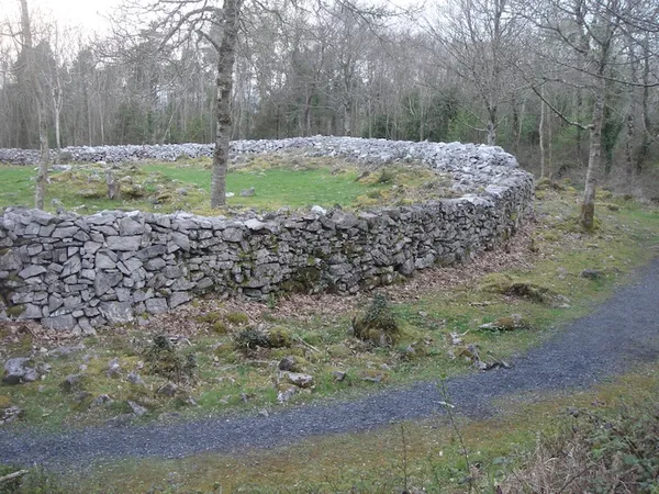 Un muro curvo in pietra a secco di calcare che racchiude un'area erbosa tra alberi spogli, con un sentiero di ghiaia intorno.