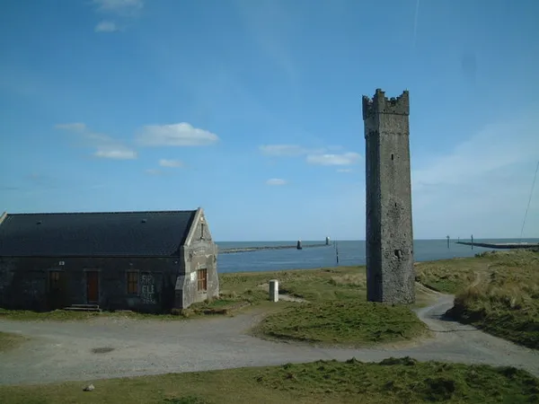 A tall slender stone tower and a low stone boathouse on a grassy shore by the sea under a blue sky.