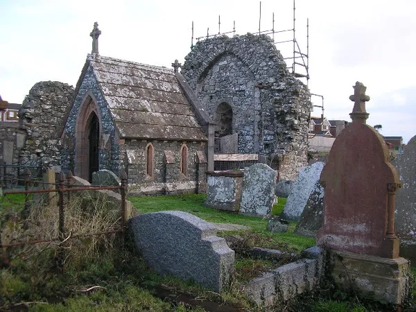Un muro de iglesia de piedra en ruinas junto a una pequeña capilla con hastial, con lápidas erosionadas entre la hierba alta.