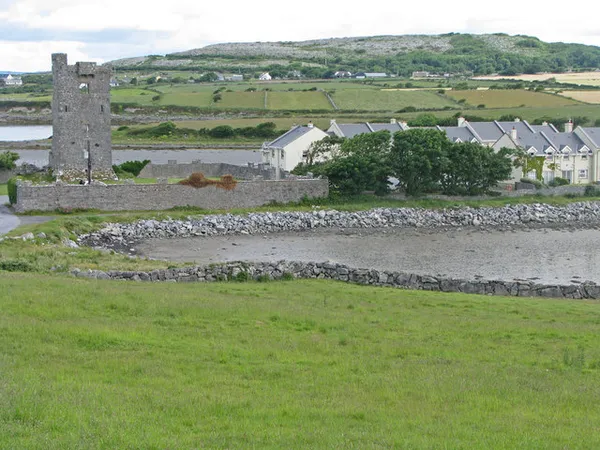 Une maison-tour en pierre en ruine sur la rive d'un chenal de marée, avec des maisons plus récentes et des champs verts derrière.