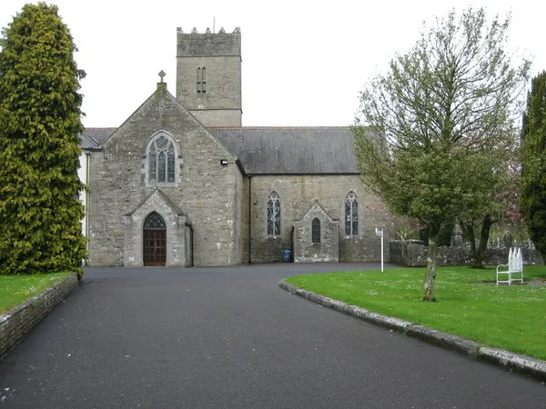A grey stone church with a square tower and Gothic windows at the end of a curving driveway and lawn.