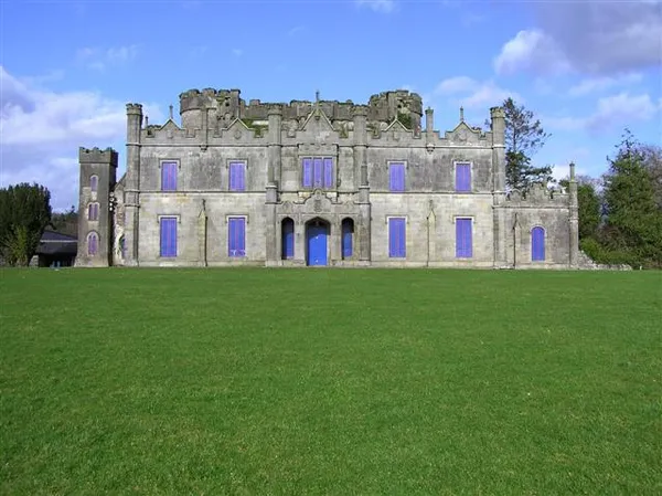 The grey stone facade of a large Gothic castle with blue-painted windows and doors behind a wide green lawn.