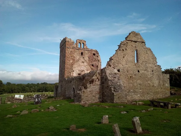 A roofless stone church with a tower standing in a grassy graveyard under a blue sky.
