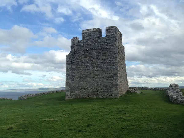 Una torre di pietra quadrata in rovina su una collina erbosa sotto un cielo nuvoloso, con il mare dietro.