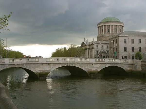 A three-arched stone bridge crossing a river in front of a domed classical building under a cloudy sky.