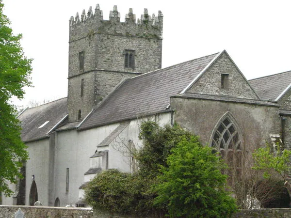 Une église en pierre grise avec une tour carrée crénelée et une fenêtre gothique en pointe, partiellement masquée par des arbres.