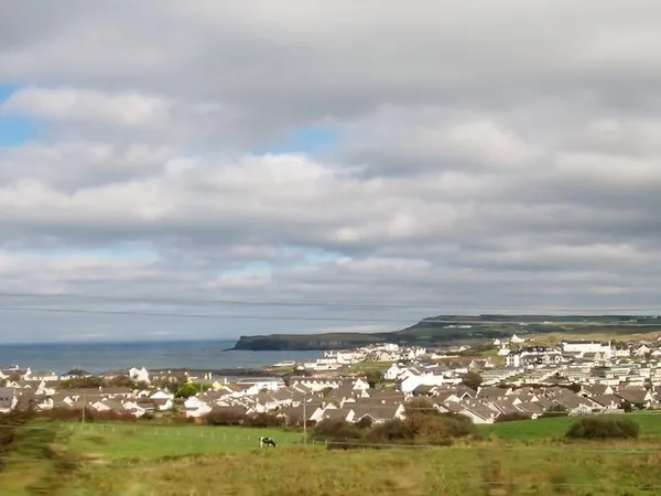 White houses of a coastal village spread between green fields and the sea, with a headland beyond.