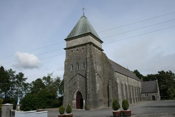 A grey stone Gothic church with a steep green roof on its tower, beside a road under a cloudy sky.