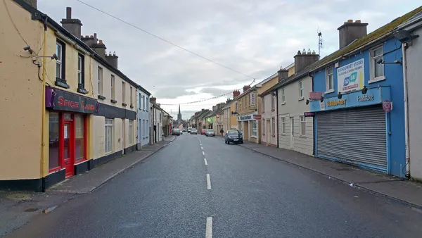 A long straight town street lined with painted shopfronts, with a church spire in the distance.