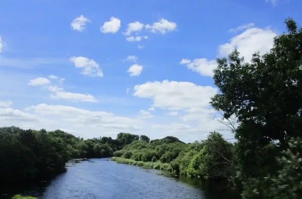 Il fiume Lee che scorre tra rive verdi boscose sotto un cielo azzurro con nuvole sparse.