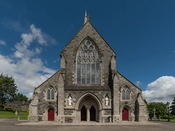 Stone Gothic cathedral front with a large traceried window and three arched red doors under blue sky.