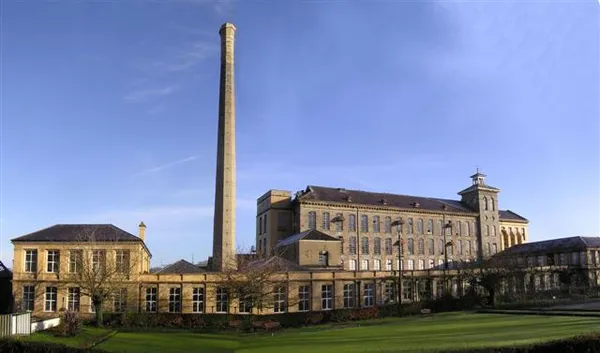 Ein großes steinernes Mühlengebäude mit einem hohen Ziegelschornstein hinter einem grünen Rasen unter blauem Himmel.