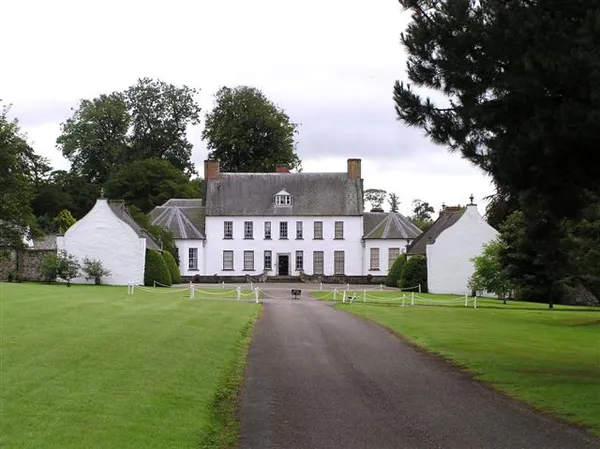 A white house with a slate roof and white side wings at the end of a drive between green lawns.