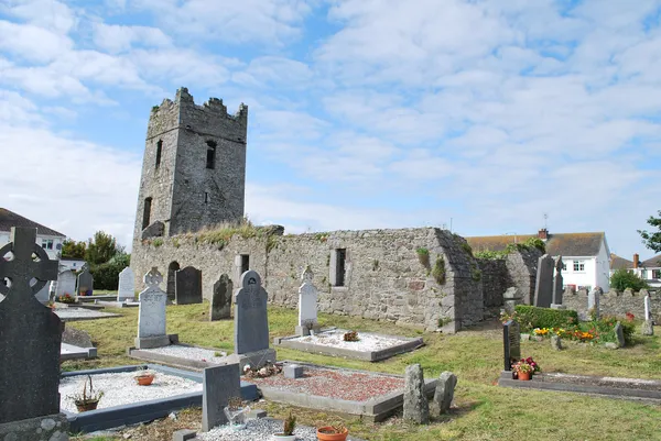 Una chiesa di pietra in rovina con una torre merlata tra lapidi sotto un cielo azzurro con nuvole.