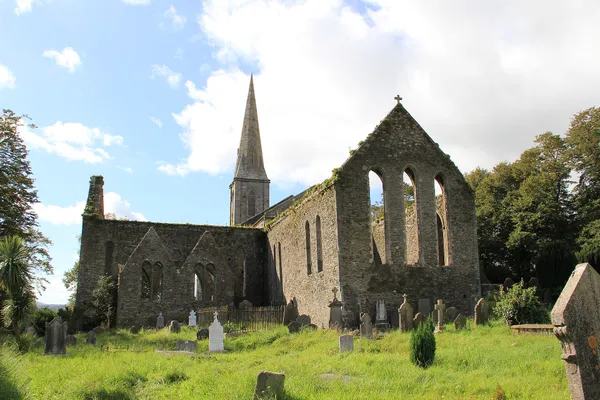 A roofless stone church with tall lancet windows in a grassy graveyard, a spire behind.
