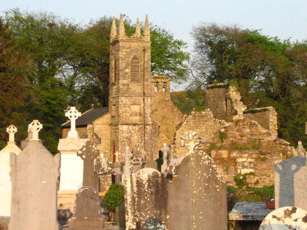 Una torre di chiesa in pietra in rovina tra vecchie rovine, con croci celtiche e lapidi davanti.