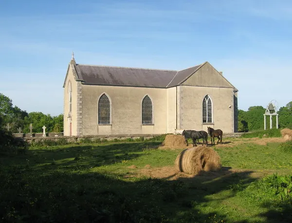 Une église en pierre pâle avec des fenêtres en arc à côté d'un champ avec des chevaux et une botte de foin ronde.
