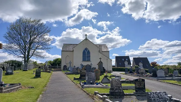 Una chiesa bianca con frontone e una croce in cima, oltre un sentiero del cimitero sotto un cielo azzurro nuvoloso.