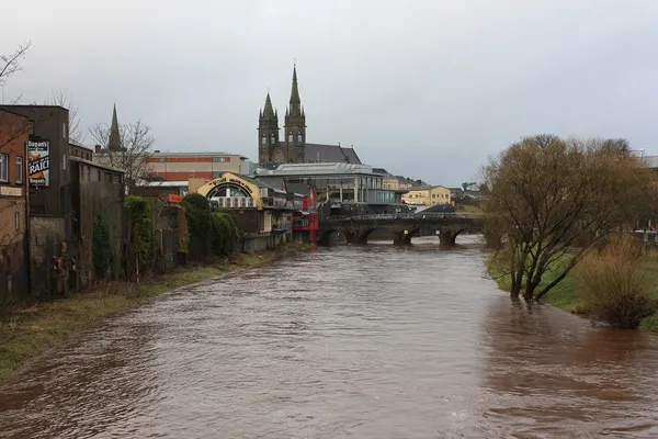Un fiume marrone che scorre attraverso una città verso un ponte, con due guglie gemelle di chiesa all'orizzonte.
