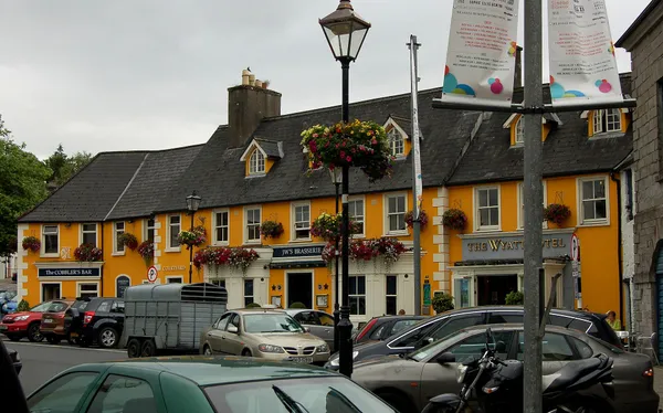 A long yellow hotel building with hanging flower baskets and a bar sign, with parked cars in front.