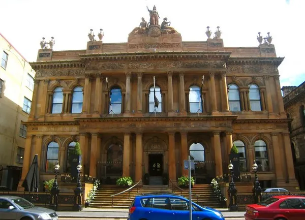 Ornate sandstone facade of a Victorian building with statues on the roofline and cars parked in front.