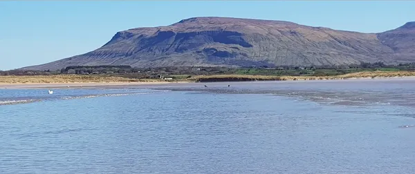 A long flat-topped mountain rising beyond calm shallow water and a low shoreline under a blue sky.