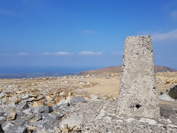A weathered concrete survey pillar on a stony summit with sea and distant hills beyond under a blue sky.